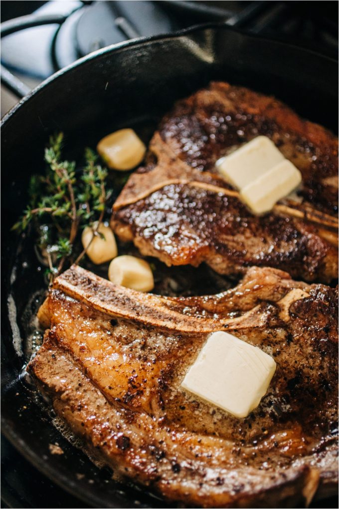 Shields Valley Ranchers rib and T-bone steaks topped with pats of butter brown nicely alongside thyme sprigs in a cast iron pan for perfect pan-seared steak on the stove.